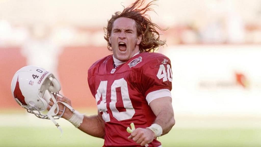 A football player celebrating on the field, wearing a red jersey with the number 40 and holding a helmet. His long hair is flowing as he cheers triumphantly.
