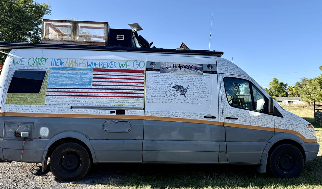 A side view of a van featuring a painted American flag and text that reads 'WE CARRY THEIR NAMES WHEREVER WE GO.' The van is decorated with names and messages honoring fallen soldiers.