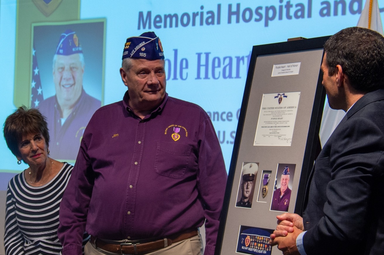 A veteran named Jim stands with a woman, smiling, as another man presents him with a framed Purple Heart certificate and photographs at a memorial event.
