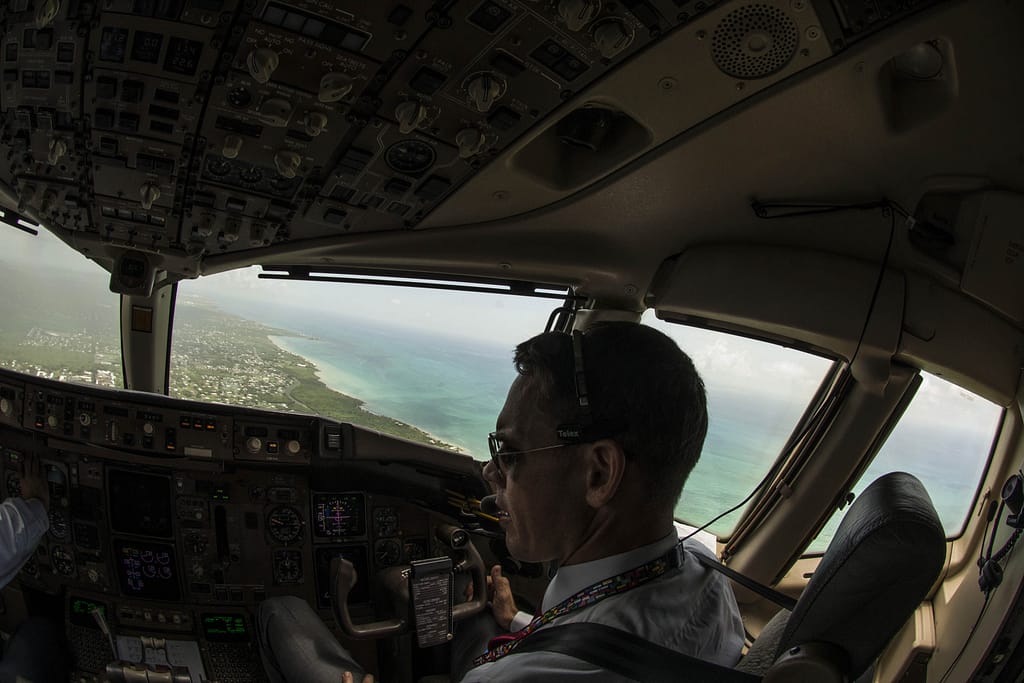 Interior view of an airplane cockpit showing a pilot operating controls with a view of the ocean and coastline in the background.