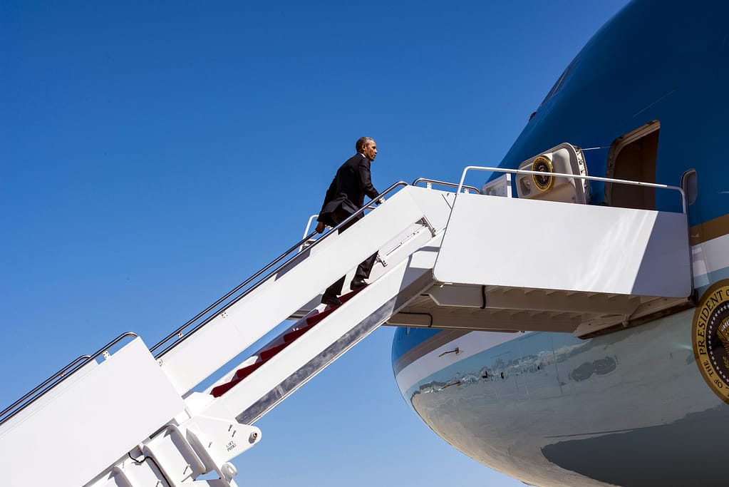 A person in a suit ascends the stairs to an airplane under a clear blue sky.