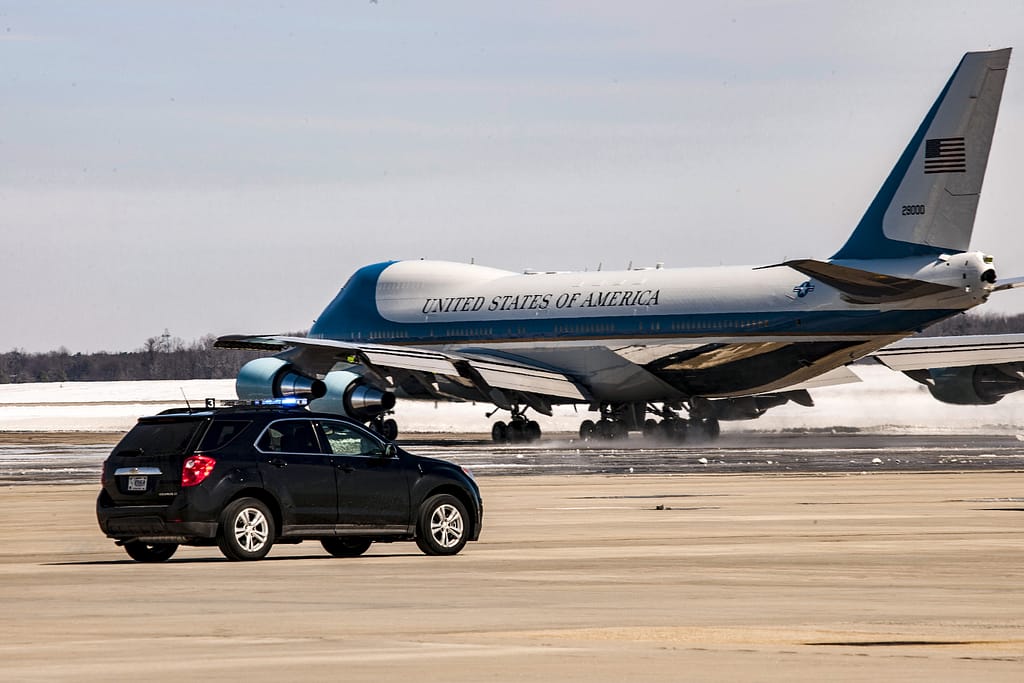 A black police vehicle with flashing lights is parked on a runway as a large aircraft marked 'United States of America' takes off in the background.