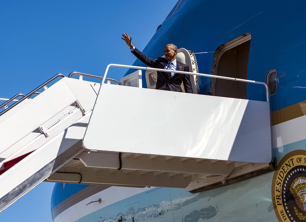 A man in a suit waves from the steps of an airplane against a clear blue sky.