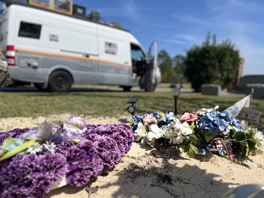 A close-up view of floral arrangements on a grave, with a white van parked in the background under a blue sky.