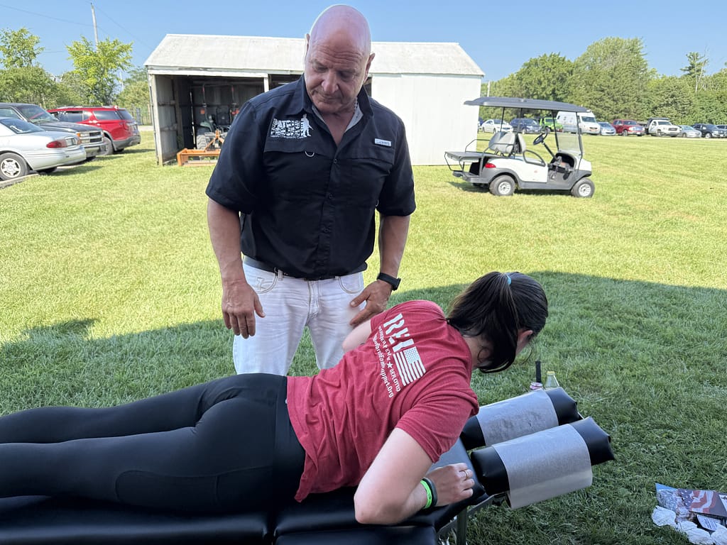 A chiropractor adjusting a person lying face down on a treatment table outdoors, surrounded by vehicles and green grass.