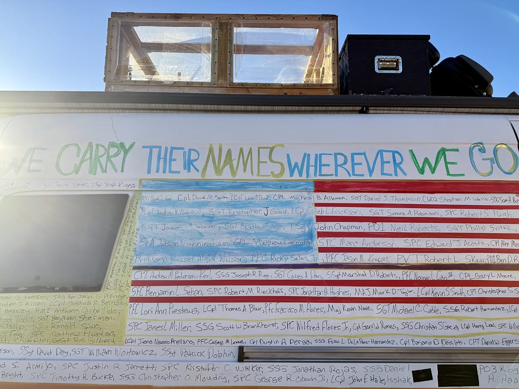 A close-up of the side of the Van of Valor featuring the words 'WE CARRY THEIR NAMES WHEREVER WE GO' along with a painted American flag and names of fallen heroes written in various colors.