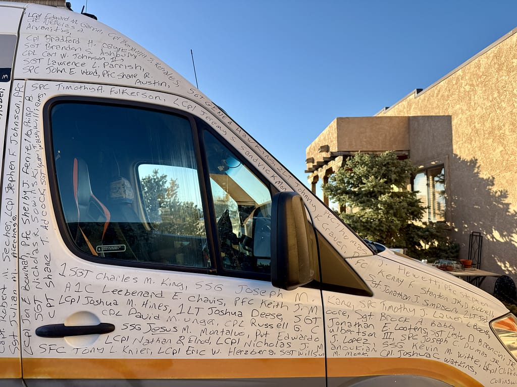 Close-up view of the side of the Van of Valor, featuring handwritten names honoring fallen heroes, with a rustic building and greenery in the background.