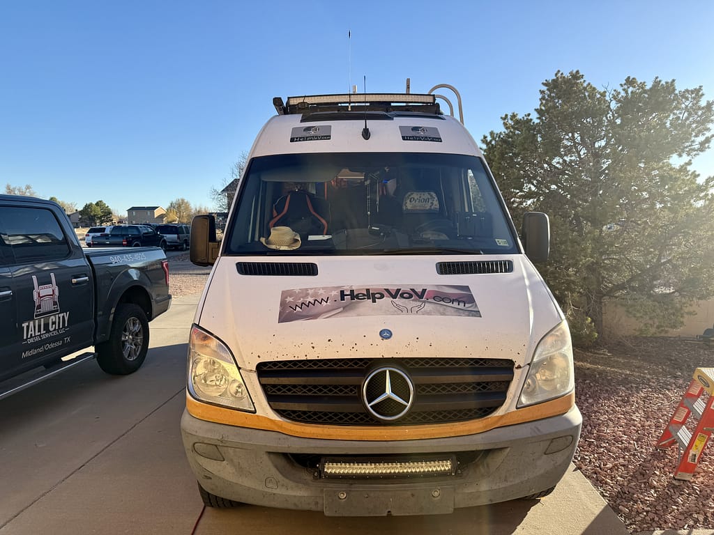 Front view of the Van of Valor, a Mercedes van with a worn exterior and a logo featuring 'HelpVoV.com,' parked in a driveway beside a pickup truck and surrounded by a landscaped yard.