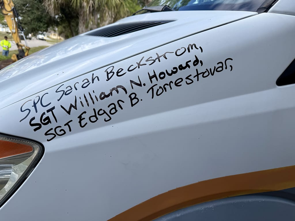 Close-up of the side of a white van displaying the names 'SPC Sarah Beckstrom', 'SGT William N. Howard', and 'SGT Edgar B. Torrestovar' inscribed in black marker.
