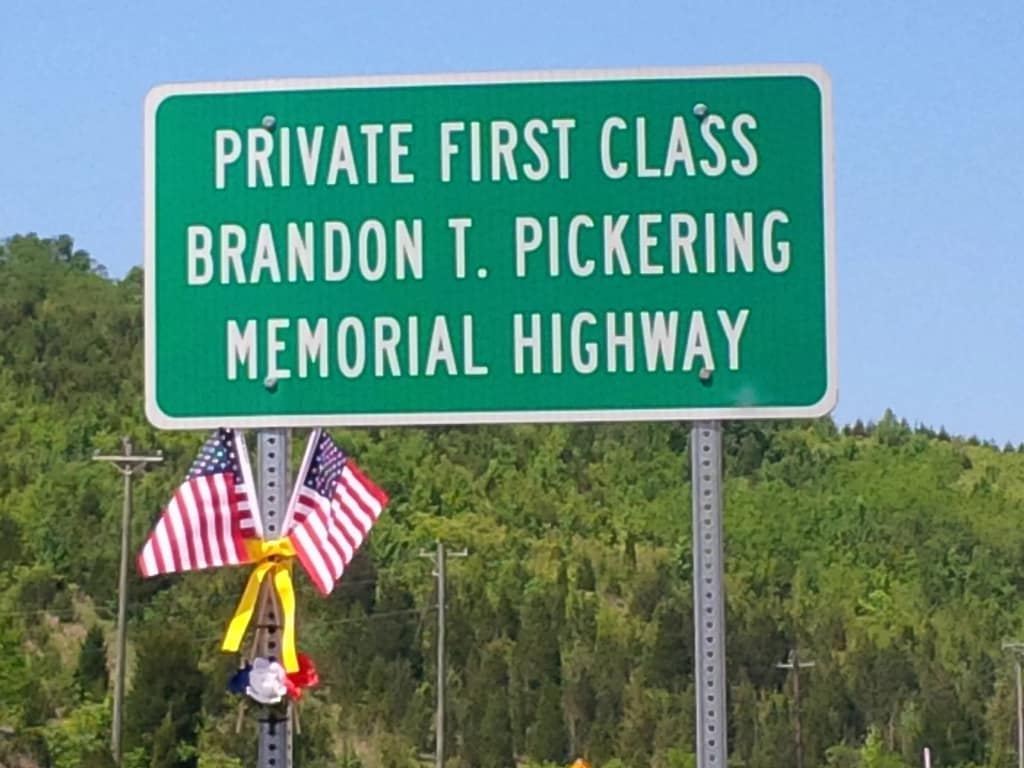Green highway sign reading 'Private First Class Brandon T. Pickering Memorial Highway' adorned with small American flags and flowers, set against a backdrop of green hills.