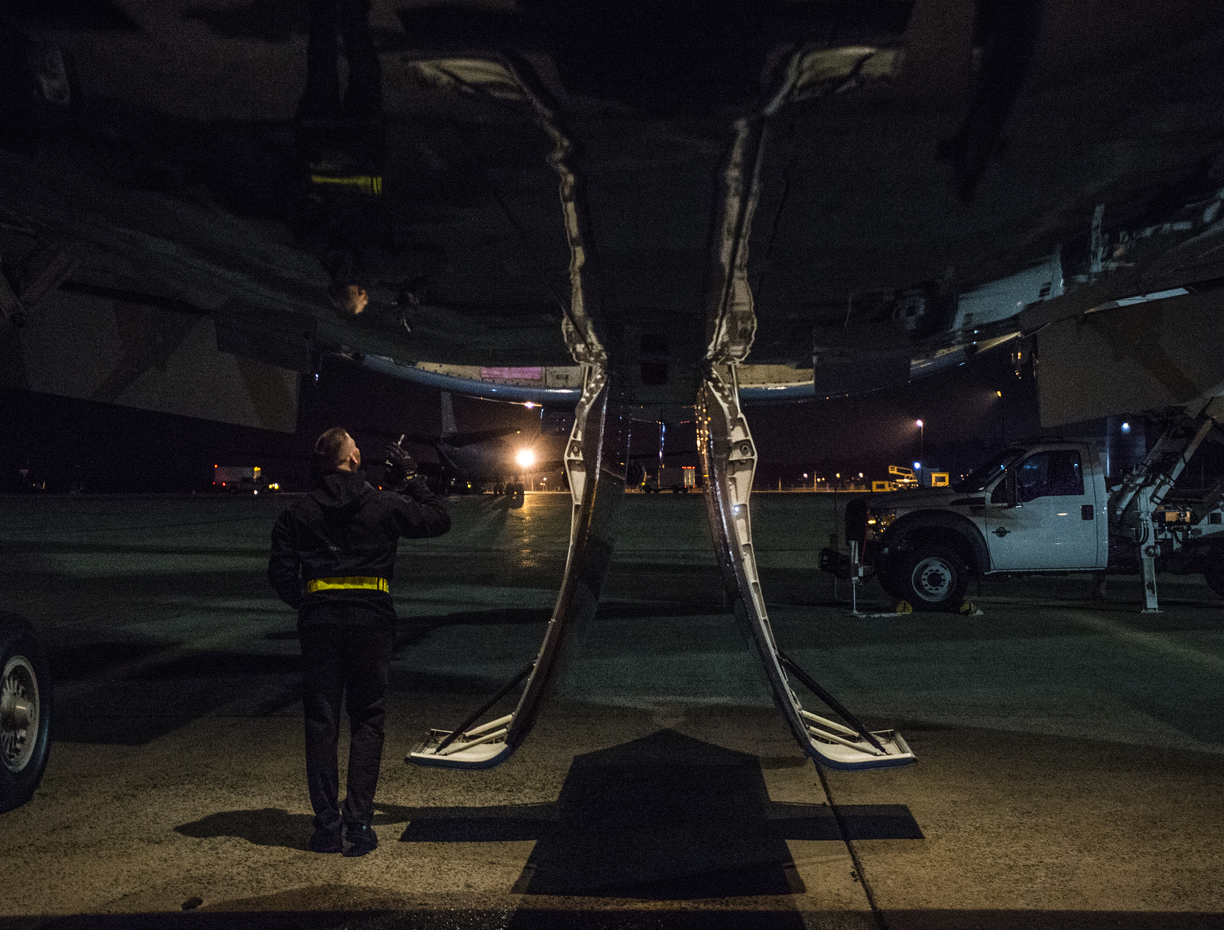 A person in a dark jacket stands in front of an aircraft on a runway, holding a camera and looking up toward the plane's wing. The scene is illuminated by soft lights in the background, creating a moody atmosphere.