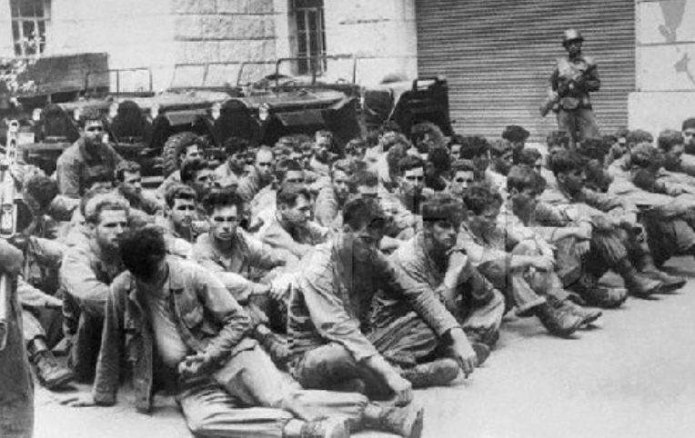 Historical black-and-white image of a group of soldiers sitting on the ground, with a soldier standing guard in the background. The scene depicts a moment of captivity, highlighting the emotional weight of their experience.