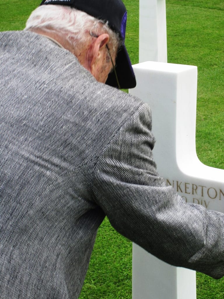 An elderly man in a gray jacket and cap gently touches the headstone of a grave at the Cambridge American Cemetery, reflecting a moment of remembrance.