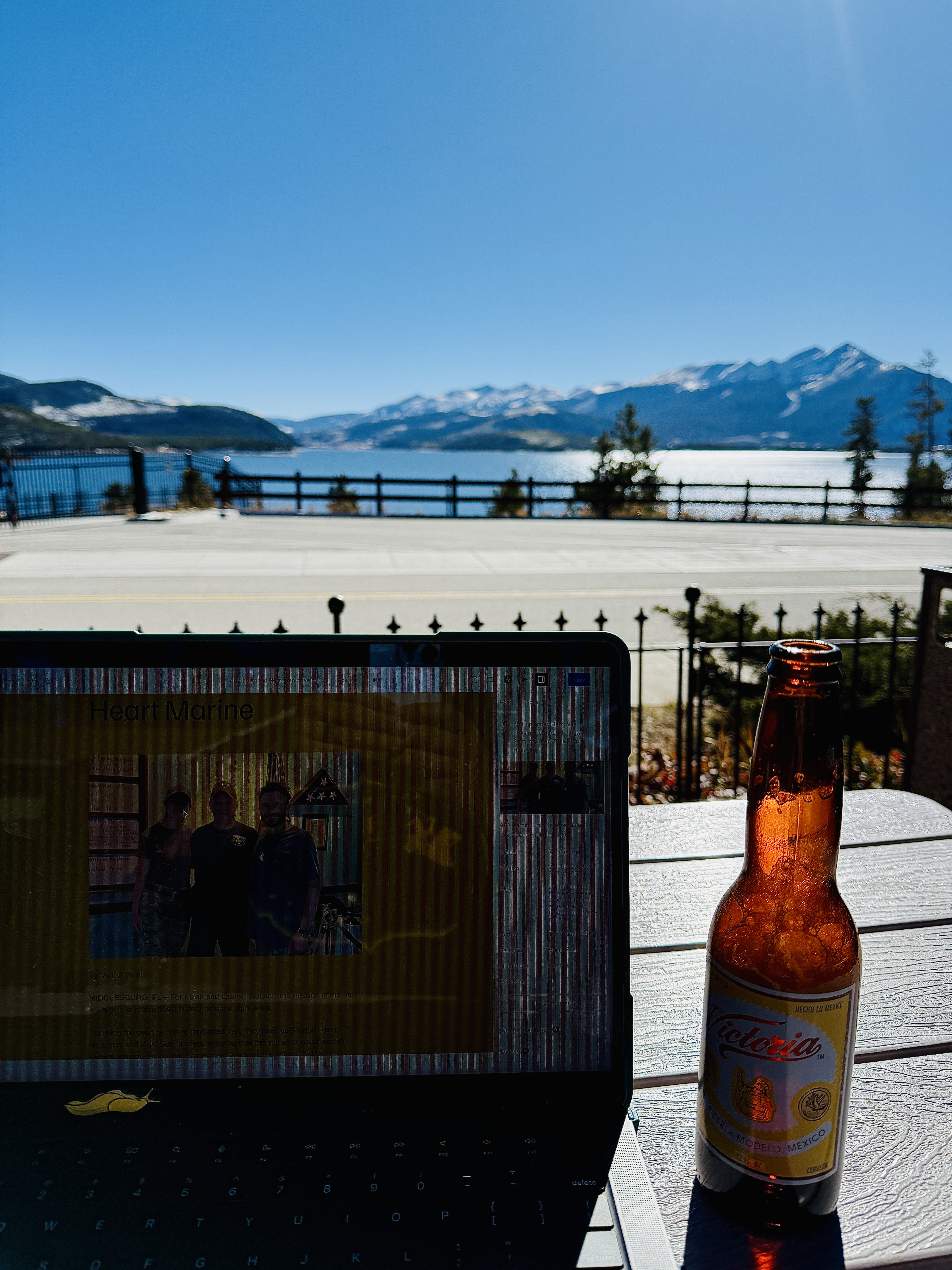 A laptop displaying a photo of three individuals in front of a mountain and lake view, with a bottle of beer on a table in the foreground under bright sunlight.