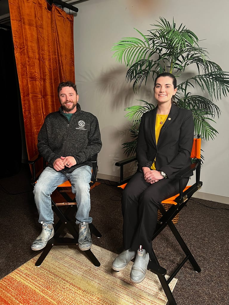A man in casual wear sits next to a woman in professional attire in front of a backdrop with plants, representing a moment of discussion or interview.
