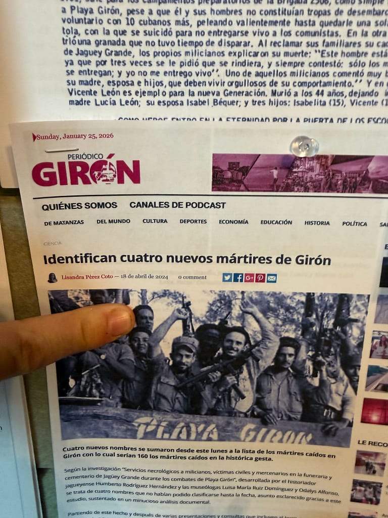 A close-up of a newspaper article titled 'Identifican cuatro nuevos mártires de Girón' from the 'Periódico Girón', detailing the identification of four new martyrs linked to the Playa Girón events, with a finger pointing to the article.